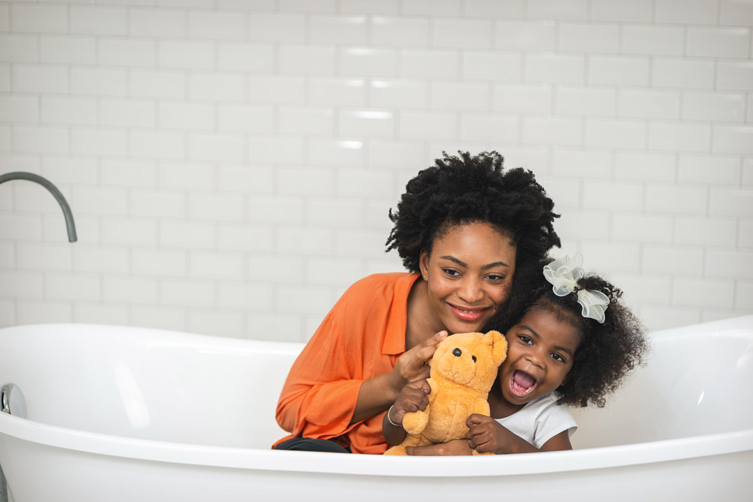 Mother and daughter clothed in white bathtub holding each other and a teddy bear