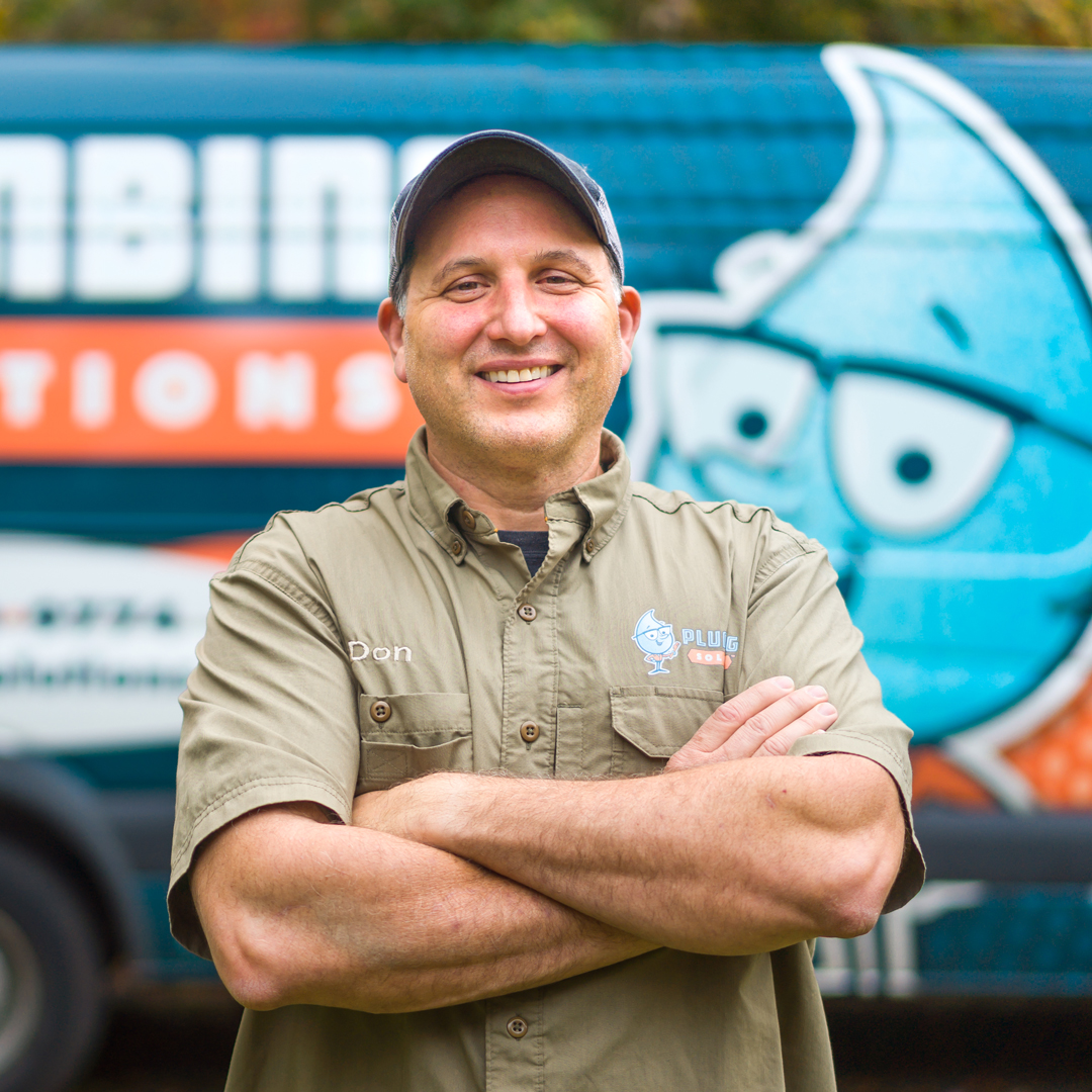 Don Meier, Owner of Plumbing Solutions Inc. stands in front of a Plumbing Solutions truck, shown from waist up with arms folded, smiling, wearing a hat