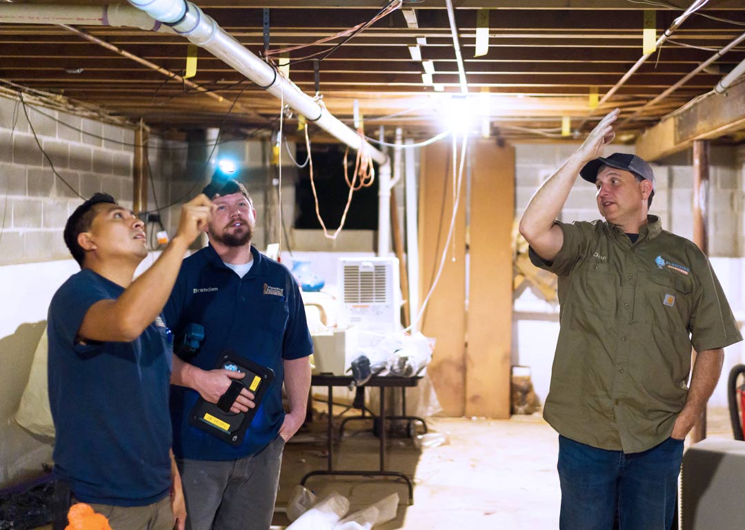 Don Meier and technicians in basement looking at floor joists and plumbing while discussing project options