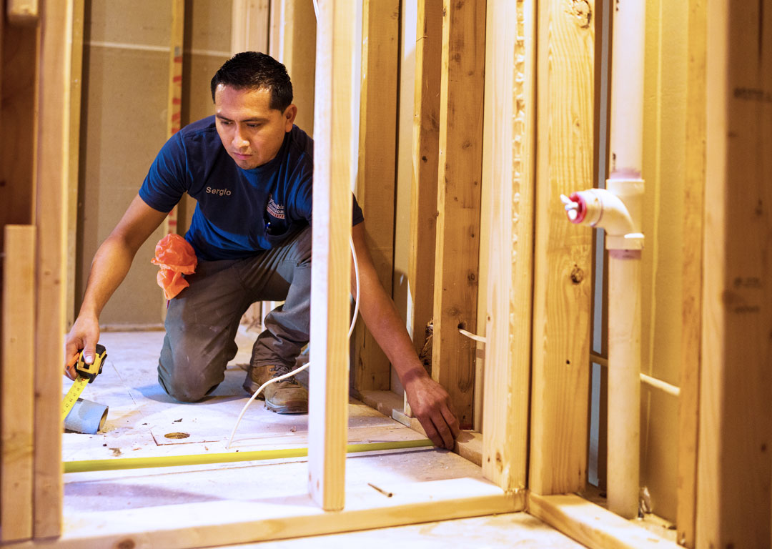 Plumbing Solutions technician kneeling in a rough framed bathroom using measuring with a tape measure